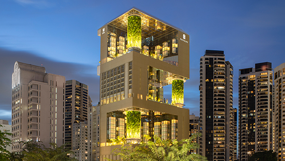 The exterior of a modern, multi-story hotel building illuminated at dusk, with glowing green architectural features and surrounding city buildings.